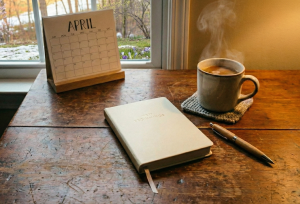 窓辺の木製テーブルに卓上カレンダー、ノート、ペン、温かい飲み物の入ったマグカップが置かれている。窓から見える外は冬から春へ移り変わる頃。