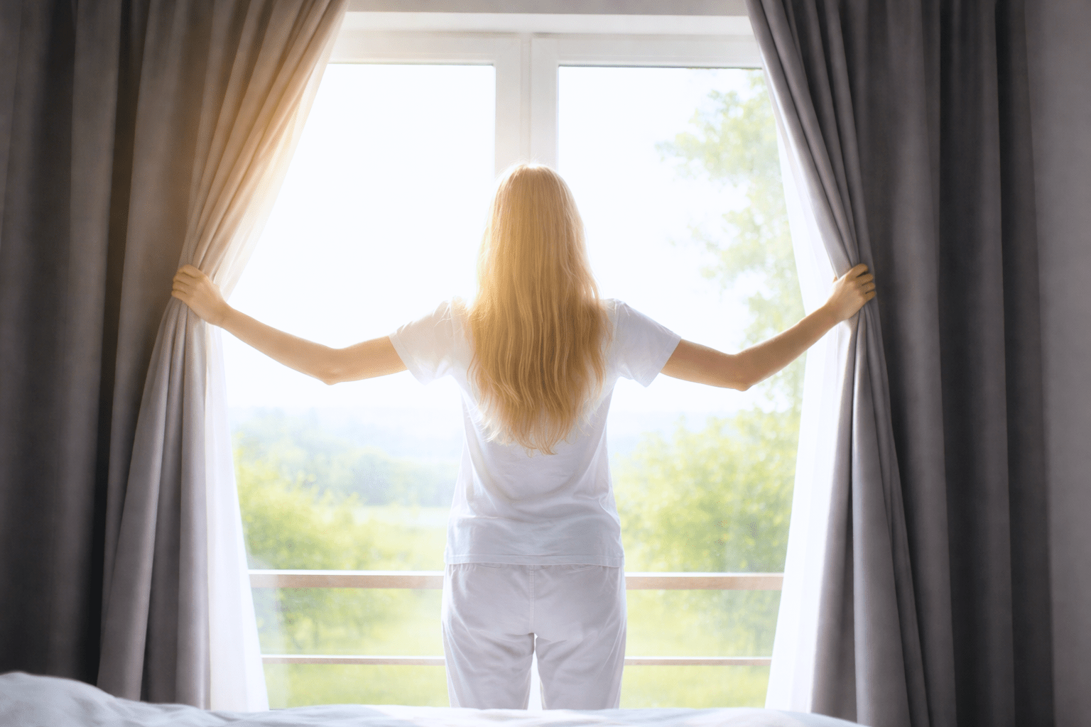 カーテンを開けて朝日を浴びる女性の後ろ姿